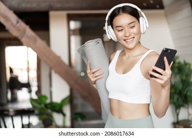 Smiling Asian Girl With Headphones, Looking At Mobile Phone And Holding Rubber Mat For Workout, Fitness At Home, Concept Of Lifestyle And Sport