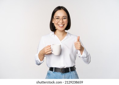 Smiling asian girl holding mug, white cup and thumbs up, recommending drink, coffee or tea, standing over white background