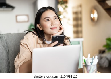 Smiling Asian Girl With Laptop And Phone, Looking Up And Thinking, Dreaming Of Something. Woman Relaxing At Home With Computer