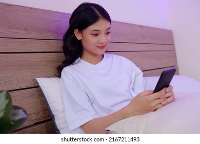 Smiling Asian Girl Lying On Bed, Checking Social Media And Setting Alarm Clock