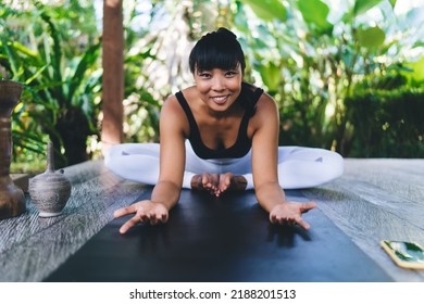 Smiling Asian Girl Practicing Yoga In Cobblers Pose And Looking At Camera. Healthy Lifestyle. Young Woman Wearing Sportswear And Barefoot On Fitness Mat On Wooden Terrace. Sunny Day On Bali Island