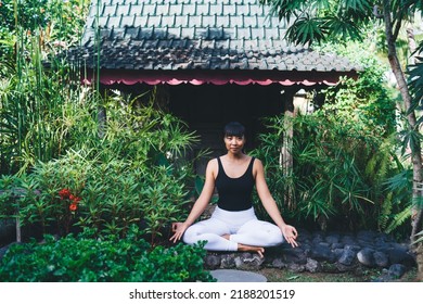 Smiling Asian Girl Practicing Yoga In Lotus Pose Outdoor. Concept Of Healthy Lifestyle. Young Woman Wearing Sportswear And Barefoot Sitting On Stones And Looking At Camera In Sunny Yard. Bali Island