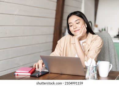 Smiling asian girl student, woman sitting with laptop at home kitchen, browing smartphone social media and resting during online classes break, waiting for webinar