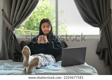 Smiling asian girl in warn clothes drinking hot chocolate and using laptop in bedroom