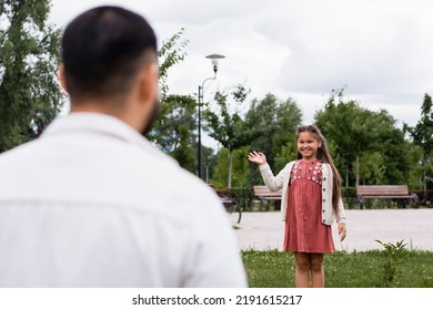 Smiling Asian Girl Waving Hand At Blurred Dad In Summer Park