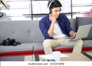 Smiling Asian handsome man sitting on sofa, he using laptop and has red headphone on his neck in living room.
