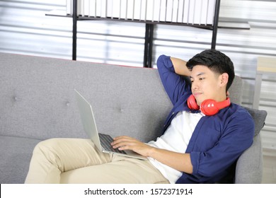 Smiling Asian handsome man sitting on sofa, he using laptop and has red headphone on his neck in living room.