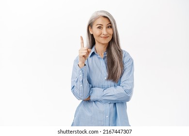 Smiling Asian Lady, Senior Woman Pointing Finger Up, Has Suggestion, Showing Promo Banner, Standing Over White Background