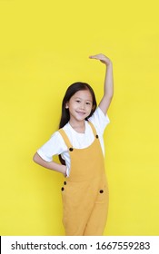 Smiling Asian Little Child Girl Measures Growth By Hand Isolated Over Yellow Background. Kid Estimate Her Height With Looking Camera