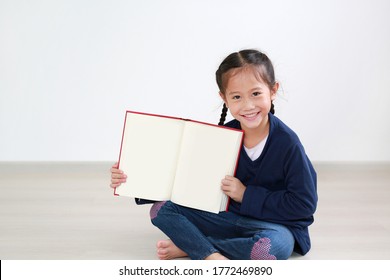 Smiling Asian Little Child Girl Open Book. Caucasian Kid Sitting In Room And Holding Blank Page Of Book.