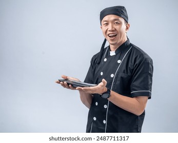 A smiling Asian male chef in a black chef's uniform and bandana, holding a stone sharpening tool with both hands, showcases his readiness and skill in the kitchen against a light blue background - Powered by Shutterstock