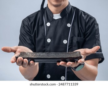 A smiling Asian male chef in a black chef's uniform and bandana, holding a stone sharpening tool with both hands, showcases his readiness and skill in the kitchen against a light blue background - Powered by Shutterstock