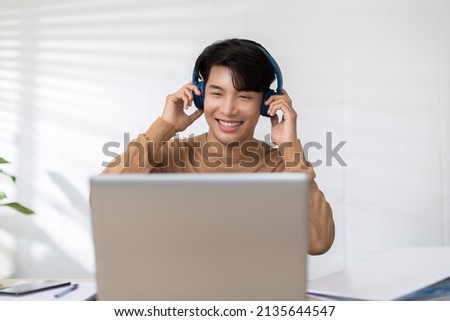 Smiling Asian male in an earphones works on laptop from home office.  Happy man in working space is talking on video call on computer. Web communication concept could helped him done a great work.