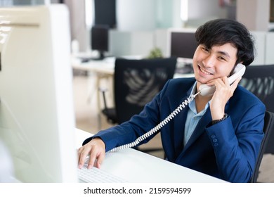 Smiling Asian Male Financial Advisor, Operator Or Telemarketer Talking On The Phone With Customer And Working With Computer At Desk In The Office. 