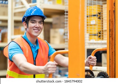 Smiling Asian Male Warehouse Worker In Safety Vest And Helmet Driving And Operating On Forklift Truck In The Industry Storage Warehouse With Shelf Pallet Automotive Spare Parts Parcel Blur Background