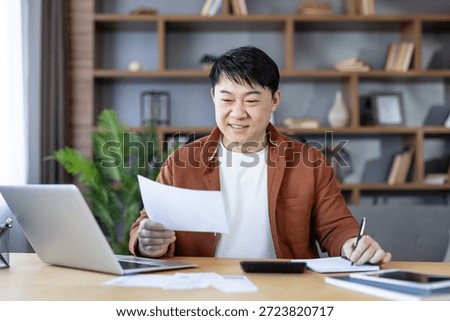 Smiling asian man analyzing documents and making notes at his laptop in a home office, proactively planning personal finances, expenses, and investment strategies