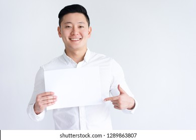 Smiling Asian Man Holding Blank Sheet Of Paper. Young Guy Pointing At Paper Sheet And Looking At Camera. Promotion Concept. Isolated Front View On White Background.