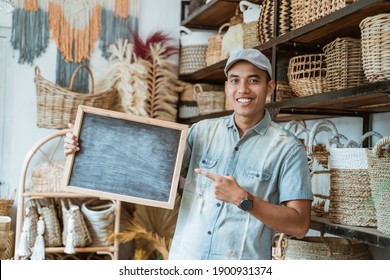 smiling Asian man looking at the camera and finger pointing to blackboard in a handicraft shop with a handicraft background