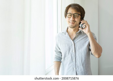 Smiling Asian Man Talking On Mobile Phone While Standing At The Window At Home.