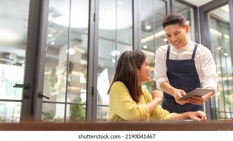 Smiling Asian Man Waiter Taking Orders From Female Client With Digital Tablet In Cafe Restaurant