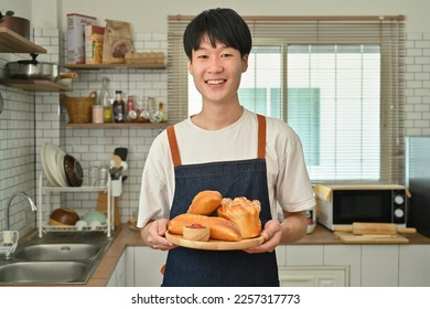 Smiling asian man wearing apron holding wooden plate with fresh homemade bread while standing in modern kitchen interior - Powered by Shutterstock