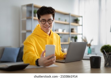 Smiling Asian Man Wearing Glasses And Yellow Hoodie Using Phone And Laptop, Working On Online Project Sitting At Desk In Modern Home Office, Young Employee Looking At Smartphone Screen, Reading Email