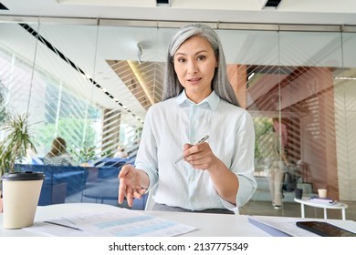 Smiling Asian Middle Aged Businesswoman Talking To Camera At Work By Videocall Conference. Financial Advisor Female Executive Consulting Client Remotely Online In Modern Office. Webcam View.