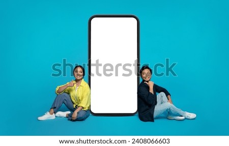 Smiling Asian millennial couple sitting on the floor with a giant smartphone between them, giving thumbs up, implying a positive review or endorsement on a turquoise background