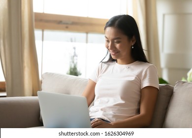 Smiling Asian Millennial Girl Sit On Couch At Home Work On Modern Laptop Gadget Texting Typing, Happy Vietnamese Young Woman Rest In Living Room Shopping Online Browsing Internet On Computer Device