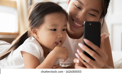 Smiling Asian millennial mom or nanny relax with little biracial girl child using cellphone together, happy Vietnamese young mother rest with small ethnic daughter watch video learning on smartphone - Powered by Shutterstock