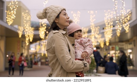 Smiling Asian Mother And Baby Turning And Looking Around At Beautiful Outdoor Christmas Lights Above Head At A Mall While Shopping For The Winter Holiday