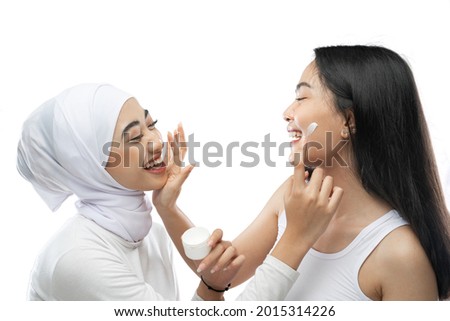 Smiling asian muslim bestfriend applying face cream with finger hands in front of camera with gray background
