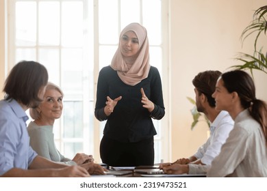 Smiling Asian muslim team leader talking to diverse employees at briefing, businesswoman wearing hijab speaking at corporate meeting, training or teaching staff, explaining project strategy - Powered by Shutterstock