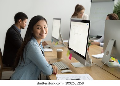 Smiling asian office employee looking at camera working with multiracial colleagues, happy young company manager sitting at corporate workplace, cheerful team member posing with desktop computer