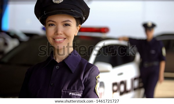 Smiling asian policewoman posing to camera against\
partner near patrol car,\
duty