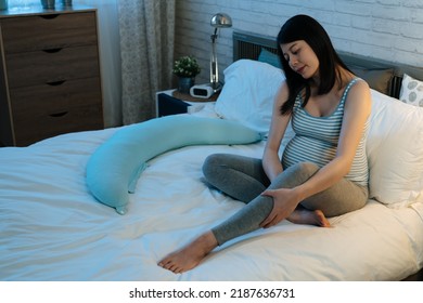 smiling asian pregnant woman sitting on bed is massaging her legs to relieve edema before going to sleep in the bedroom at home during nighttime. - Shutterstock ID 2187636731