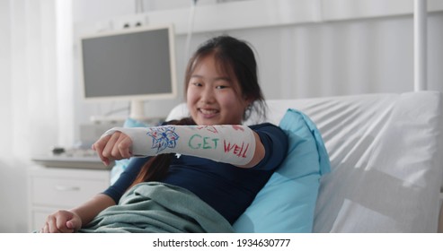 Smiling Asian Preteen Girl Lying In Hospital Bed With Broken Arm In Colored Cast. Portrait Of Chinese Kid Patient Resting In Bed With Arm In Plaster Case Painted With Colorful Markers