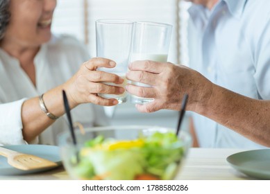 Smiling Asian Senior Man And Woman Holding Glasses Of Milk. Cheerful Elderly Couple Enjoying Healthy Salad Food While Having Breakfast At Home. Happy Lifestyle Grandfather And Grandmother.