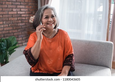 Smiling asian senior woman using phone sitting on couch at home.
