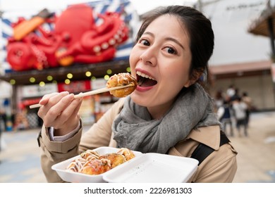 smiling Asian Taiwanese female tourist looking at camera while enjoying eating yummy takoyaki octopus ball on chopsticks in Shinsekai area in Osaka Japan