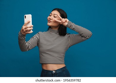 Smiling asian teen girl gesturing while taking selfie on cellphone isolated over blue background