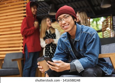 Smiling asian teenager sitting outdoors, holding takeaway coffee cup, using mobile phone - Powered by Shutterstock