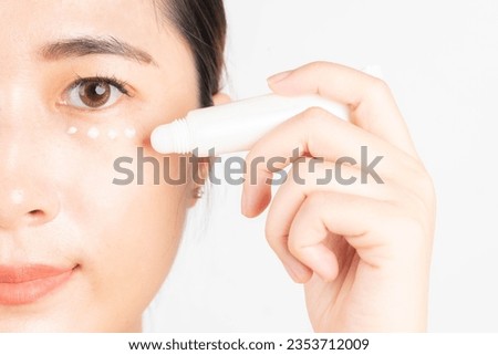 Smiling asian woman appying eye cream tube for dark circles treatment and anti aging. Skin care routine and cosmetology concept. Close up.