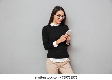 Smiling asian woman in business clothes and eyeglasses writing message on smartphone over gray background