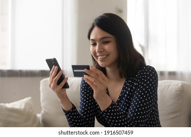 Smiling asian woman capable ebanking app user hold credit card smartphone satisfied with easy quick safe way to pay. Positive young lady sit on couch check bank account balance provide distant payment