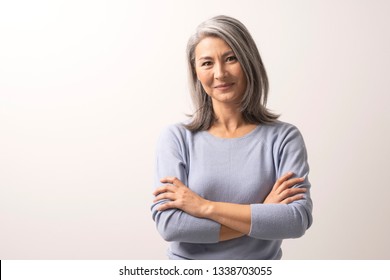 Smiling Asian Woman With Crossed Hands Stands In Front Of White Background. Charming Grey-Haired Woman With Crossed Arms Gently Smiles. Portrait.
