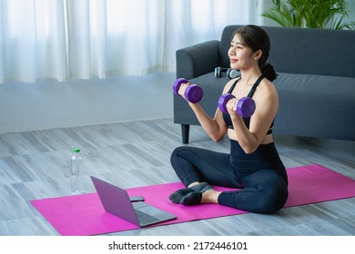 Smiling Asian Woman Doing Shoulder Stretching Yoga Online Class From Laptop At Home In Living Room. Self-isolation And Exercise At Home With A Sofa And Laptop Next To It.