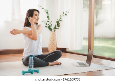 Smiling Asian Woman Doing Yoga Shoulder Stretching Online Class From Laptop At Home In Living Room. Self Isolation And Workout At Home During COVID-19.