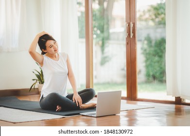 Smiling Asian Woman Doing Yoga Neck Stretching Online Class From Laptop At Home In Living Room. Self Isolation And Workout At Home During COVID-19.