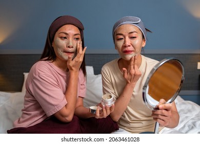 Smiling Asian Woman Friends Sitting On The Bed With Applying Skin Care Facial Mask On Their Face Together At Home. Female Gay Couple Relax And Enjoy Beauty Facial Treatment Together With Happiness